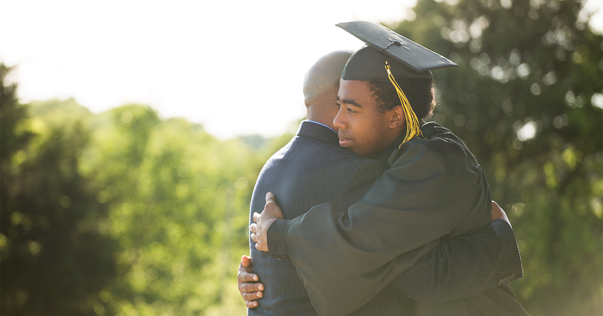 Black college graduate in cap and gown hugging his father