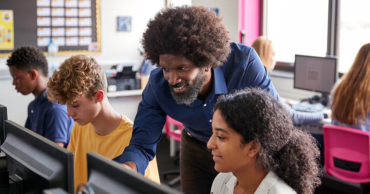 Teacher instructs high school student over her shoulder, looking at a computer together