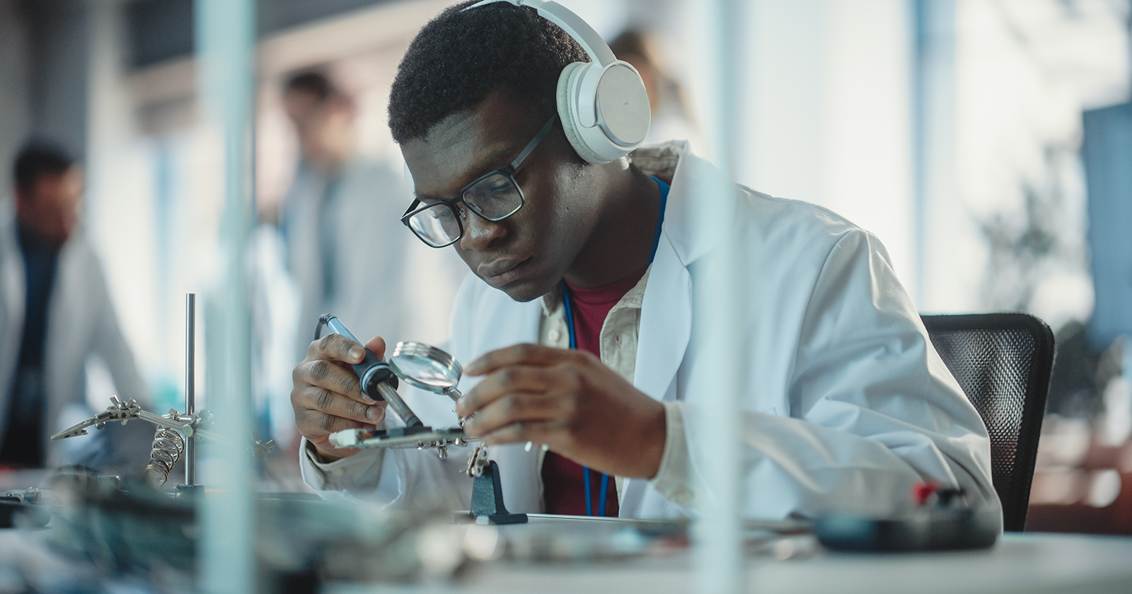 Black male student soldering a circuit board wearing headphones, a lab coat, and goggles