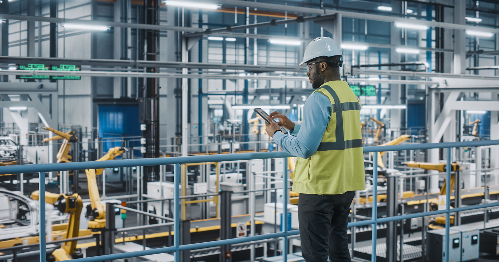 Car factory engineer in a vest and hard hat using a tablet computer and looking at the production line