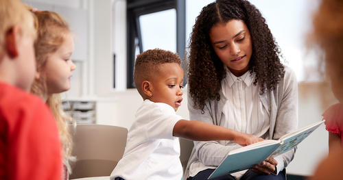 boy showing something in a book holding by teacher with other kids nearby